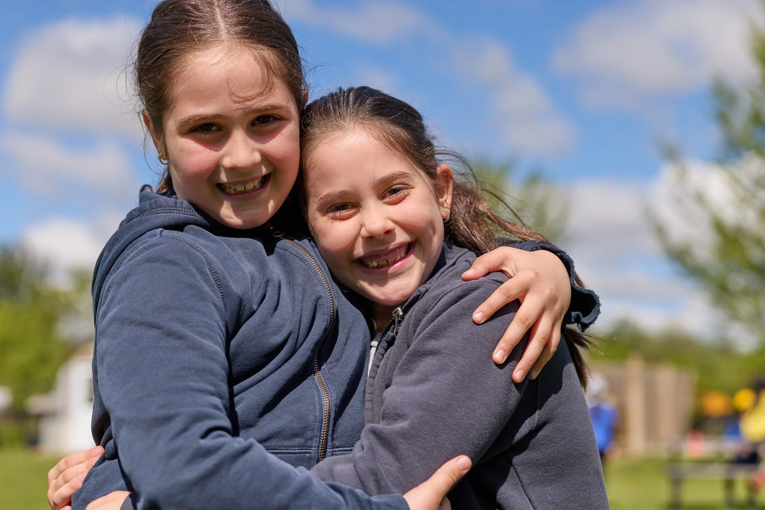 Two girls hugging outdoors on a sunny day, smiling at the camera.