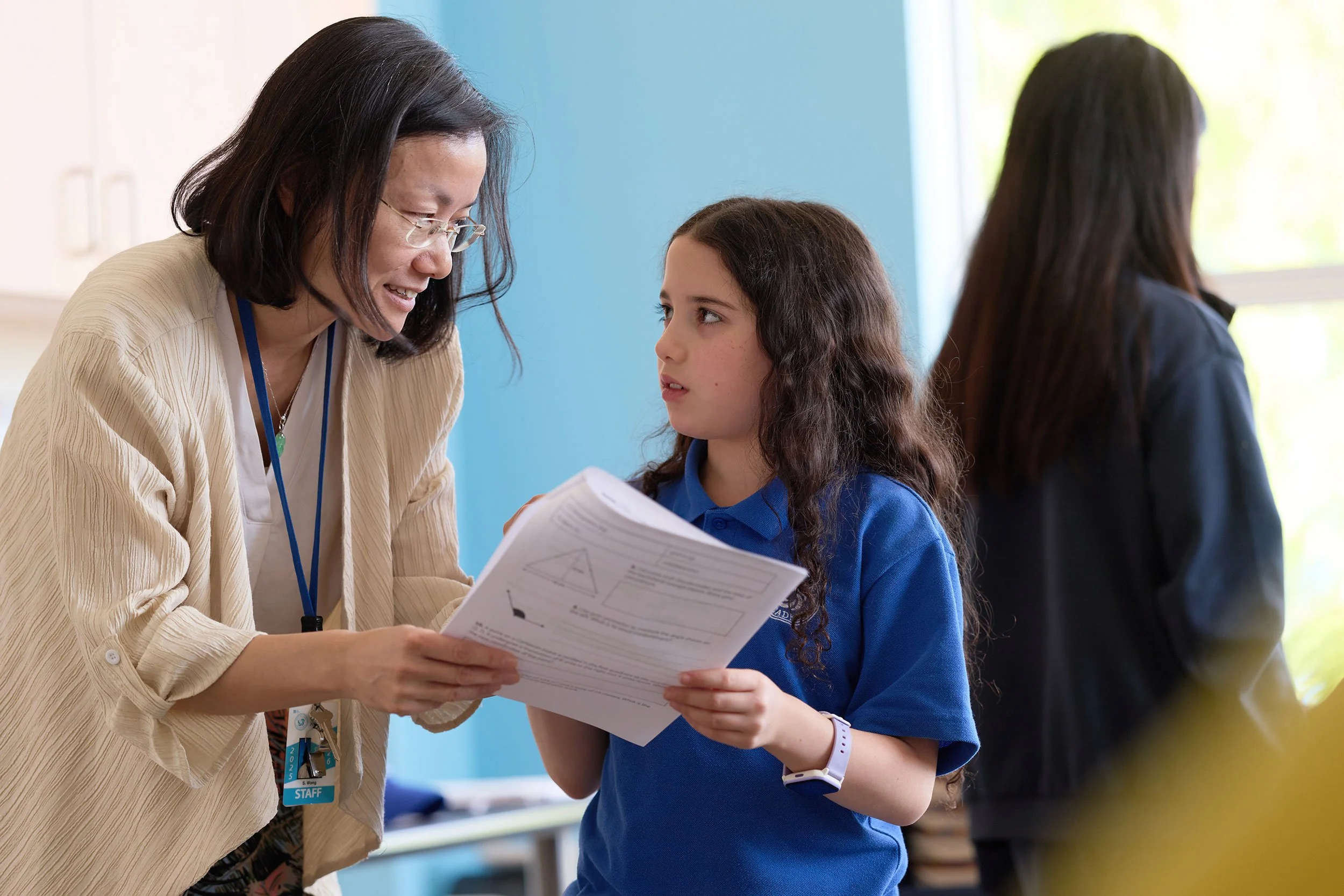 A woman with glasses and a beige blazer smiling while helping a young girl in a blue polo shirt with homework at a school setting.