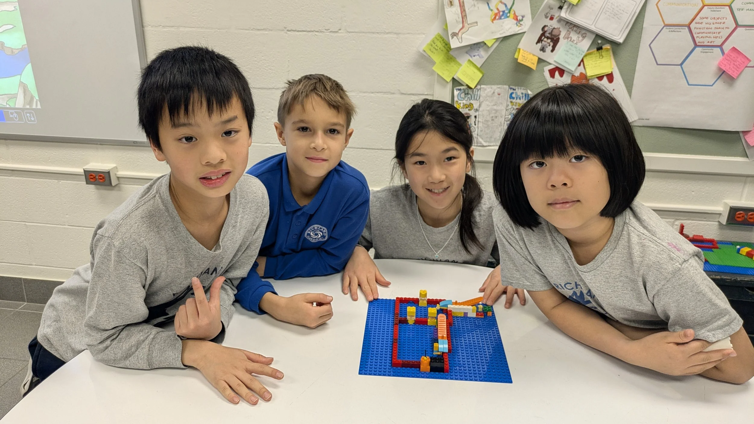 Four children sitting at a table with a blue LEGO creation in front of them, inside a classroom with colorful posters on the wall.