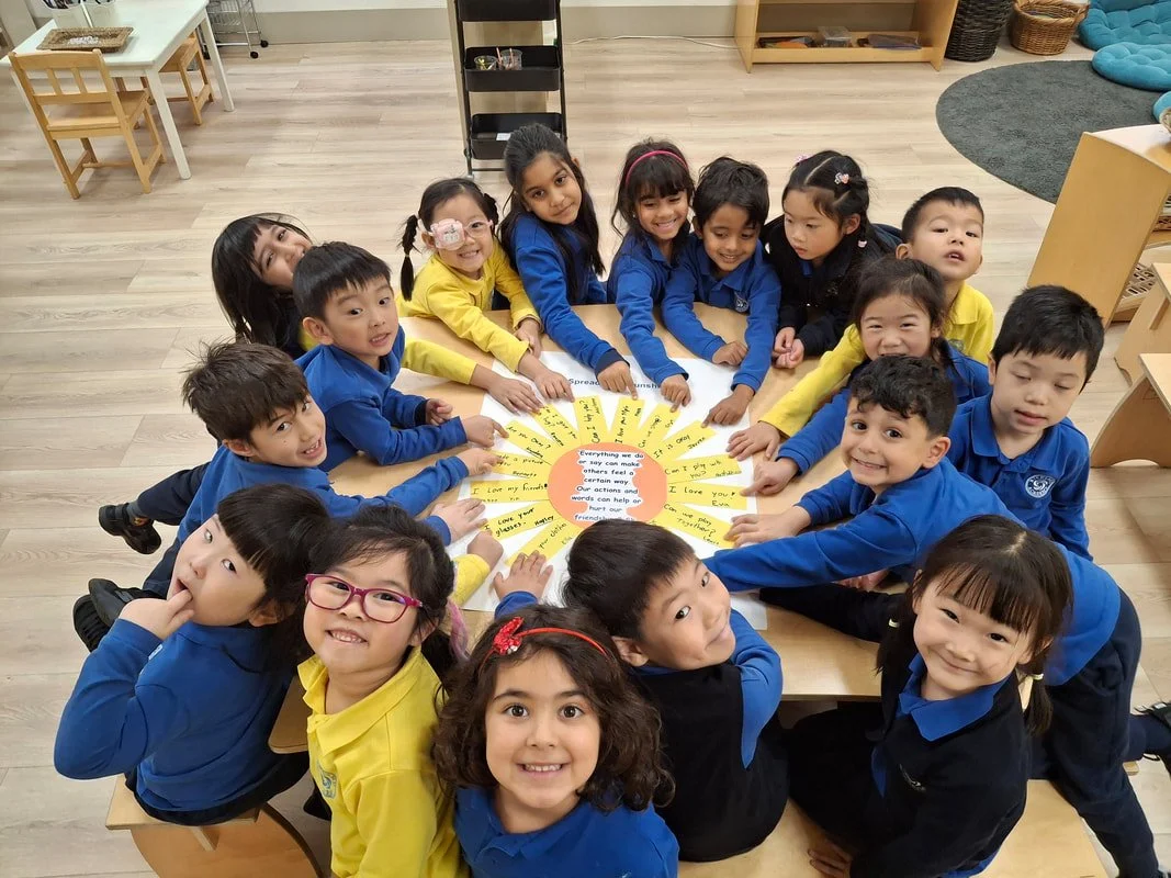 Group of young children gathered around a table in a classroom, pointing at a colorful circular chart with educational messages.