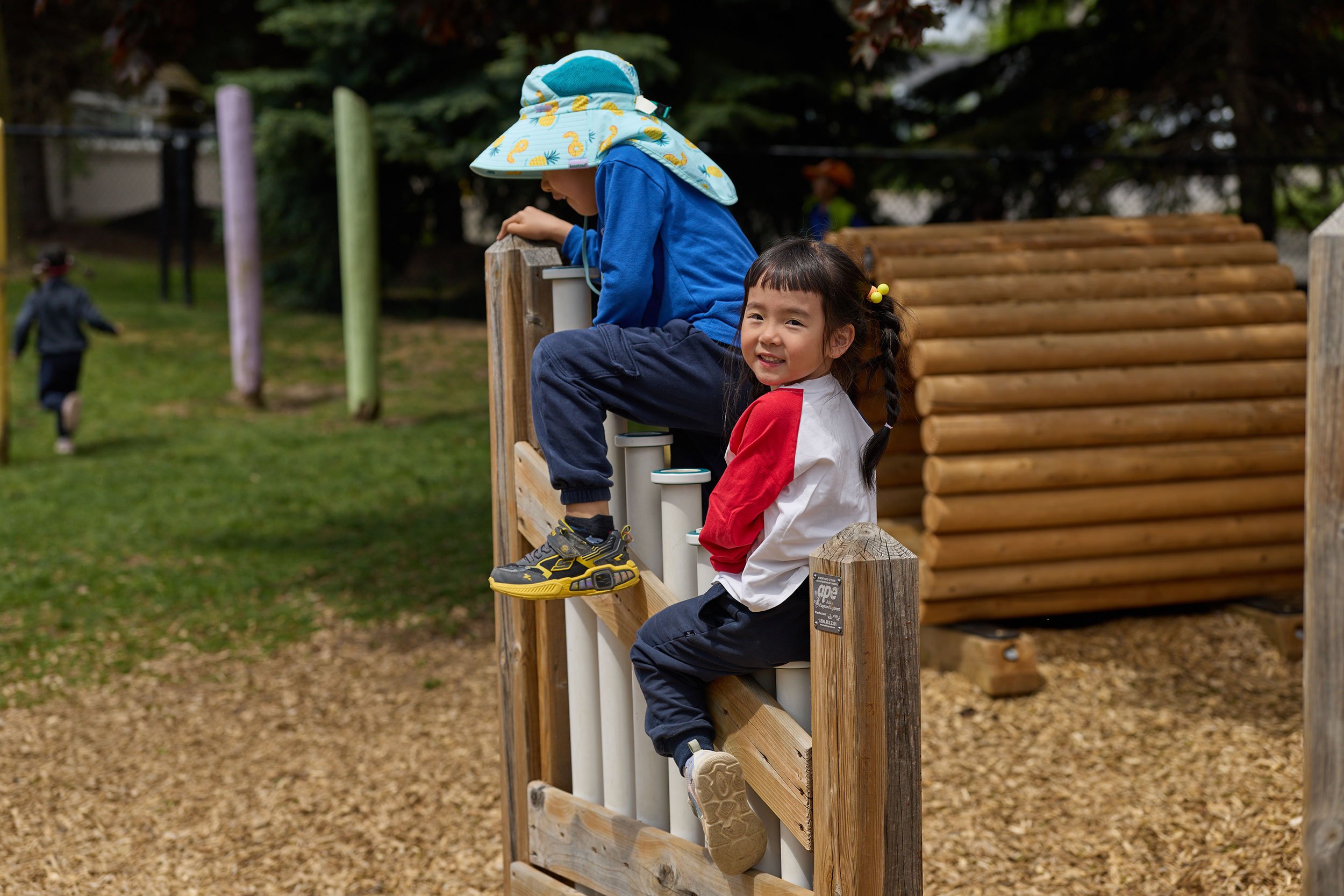 Smiling young girl with braided hair and yellow hair accessories sitting on a wooden structure at a playground, with a boy in a blue jacket and sun hat climbing on the play equipment in the background.