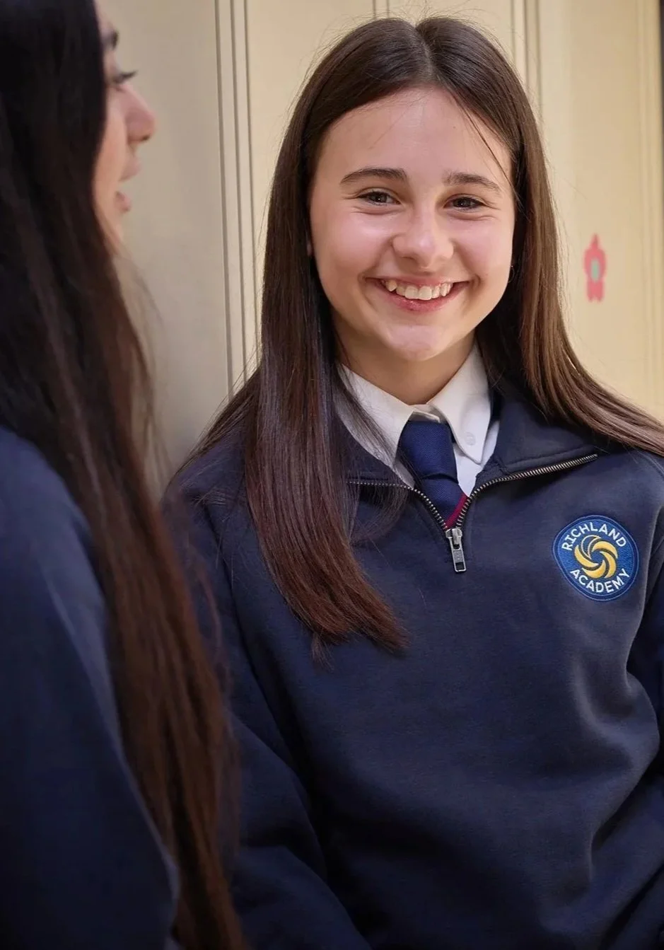 Two girls in school uniforms talking, one girl smiling at the camera, standing near lockers.