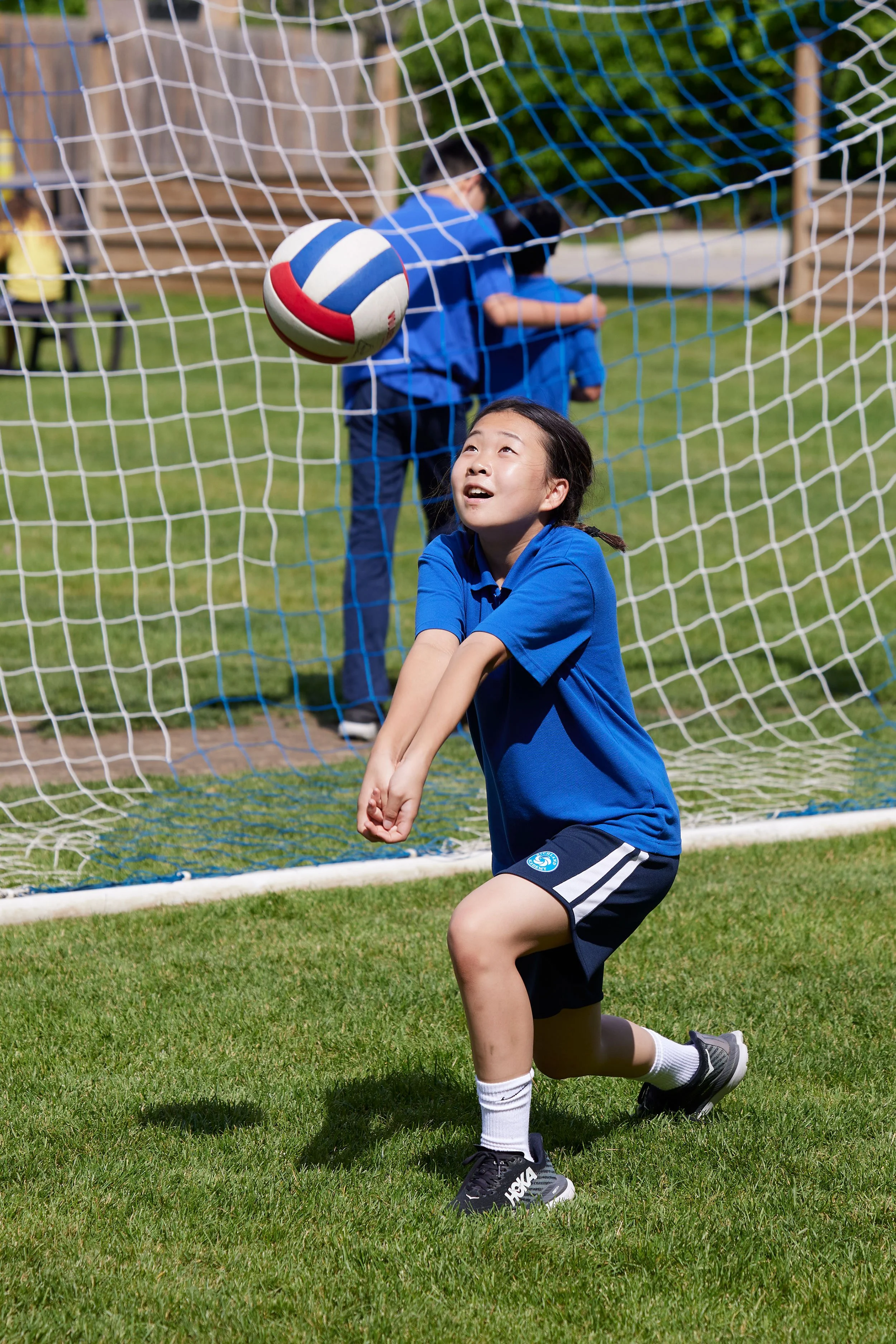 A young girl in a blue sports uniform is kneeling on the grass, looking up at a volleyball as it hits the goal net. Other children in blue uniforms watch in the background.