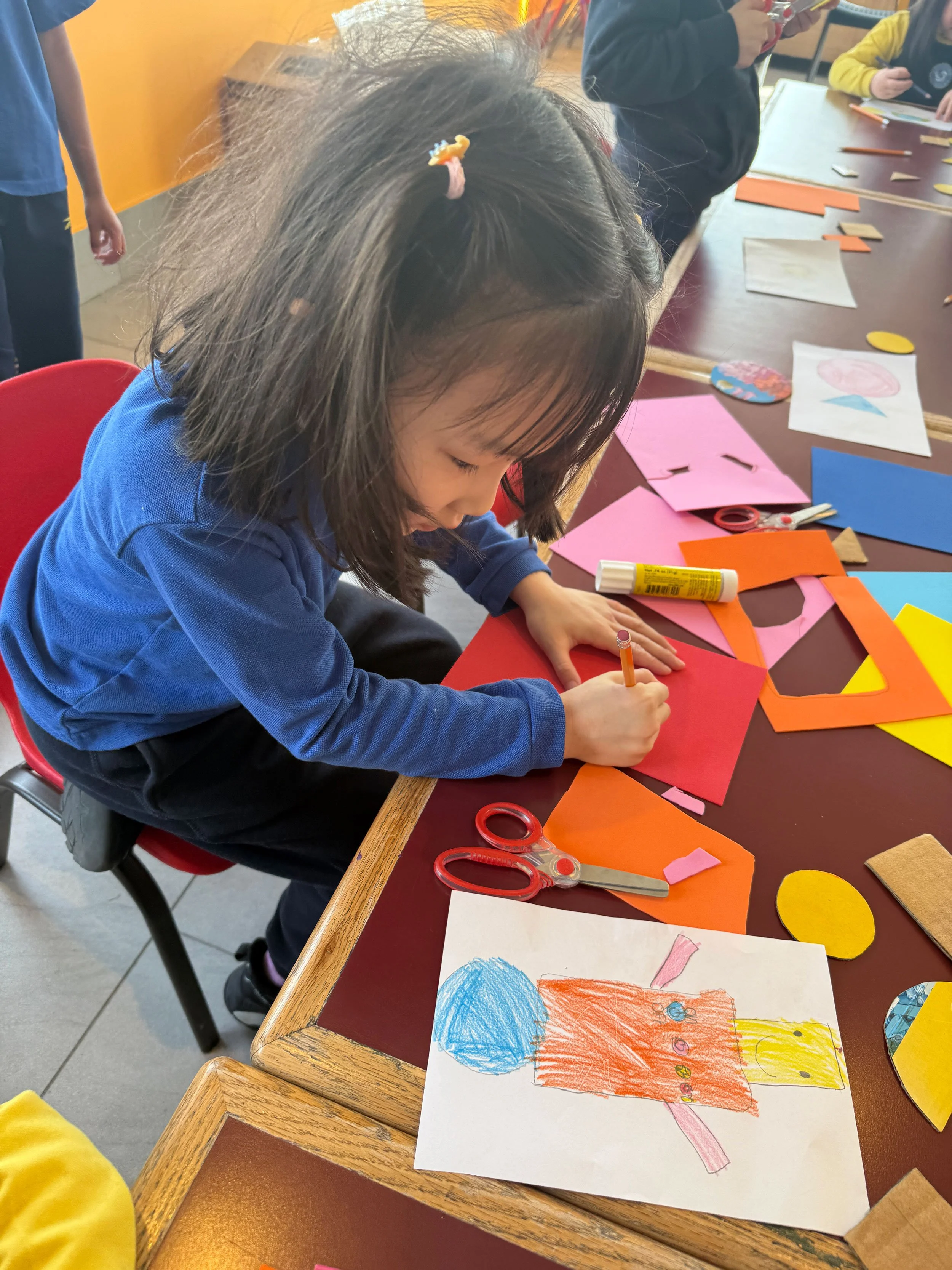 A young girl sitting at a table drawing with colored markers on paper, surrounded by craft supplies including scissors and colored paper. Other children are visible in the background working on similar projects.