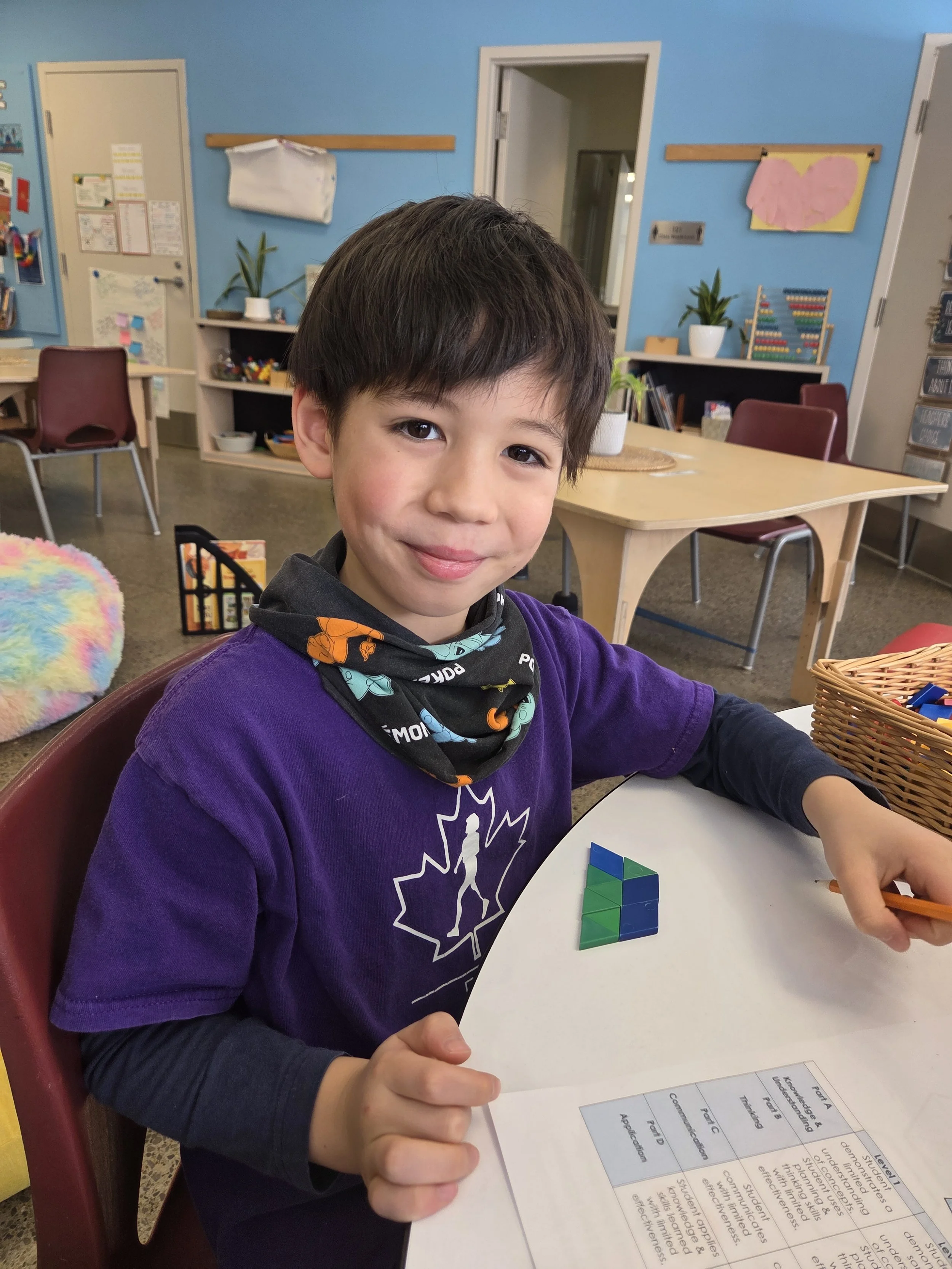 Young boy sitting at a classroom table, smiling, with a colorful plastic puzzle in front of him, wearing a purple sweatshirt and a black neck gaiter with cartoon characters, in a decorated classroom.