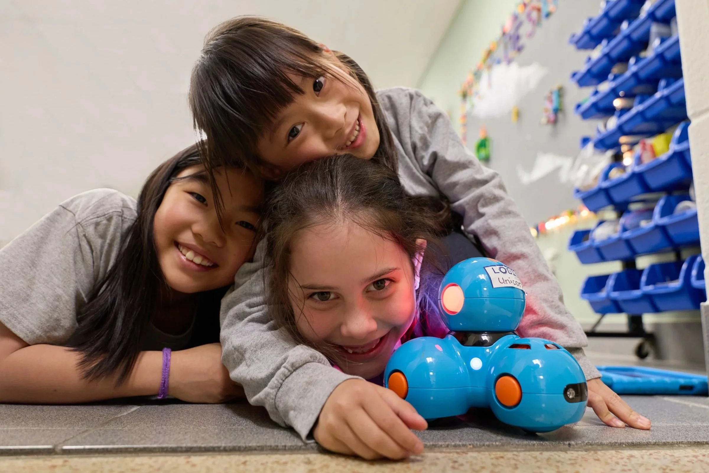 Four children are lying on the floor and playing with a blue robotic toy dog. They are smiling and seem to be having fun.
