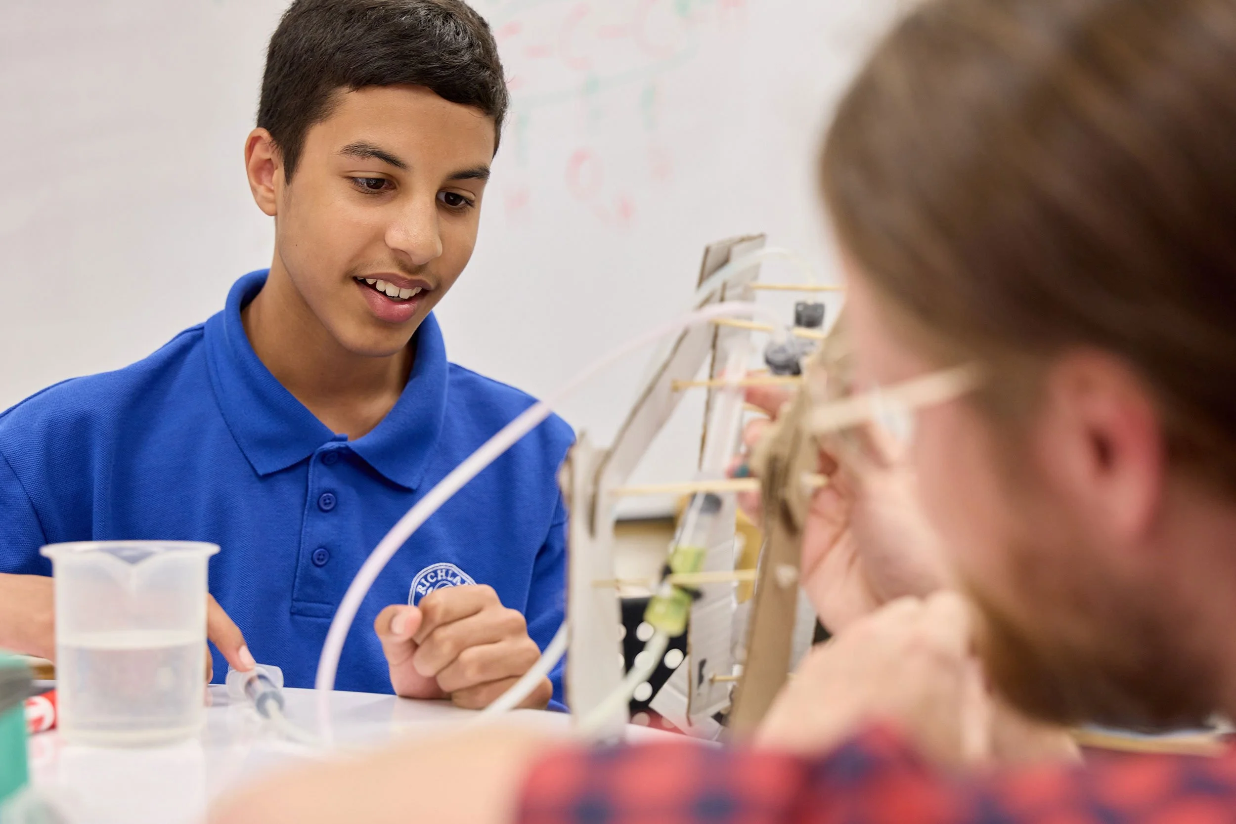 A young man wearing a blue shirt with a logo interacts with a young girl in a classroom or lab, with scientific equipment and a whiteboard in the background.