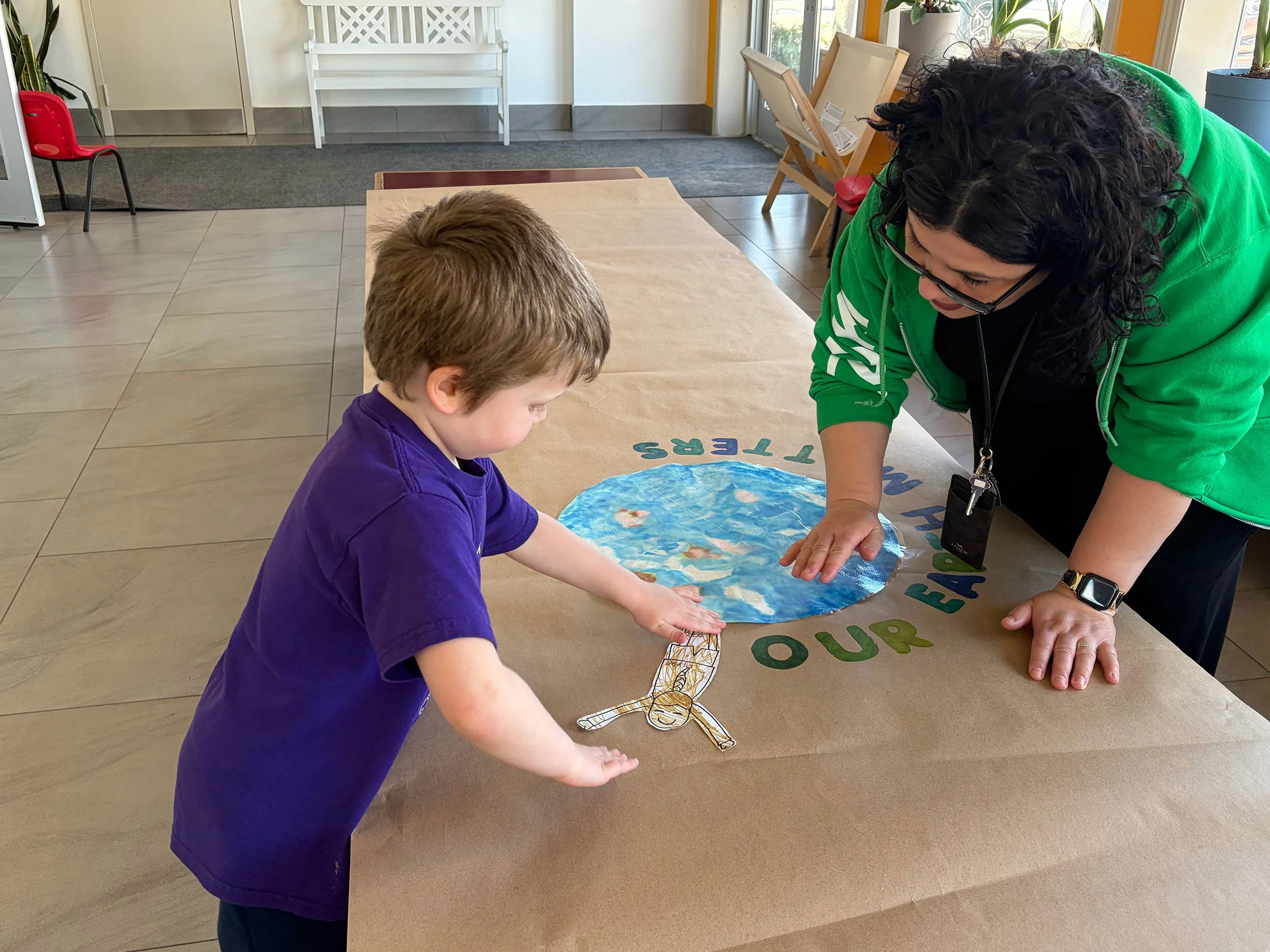 A woman with curly black hair wearing glasses and a green hoodie looks down as she helps a young boy with brown hair and a purple shirt craft a paper fish on a table with a circular paper Earth on it. The setting is a bright indoor space with chairs, plants, and a door in the background.