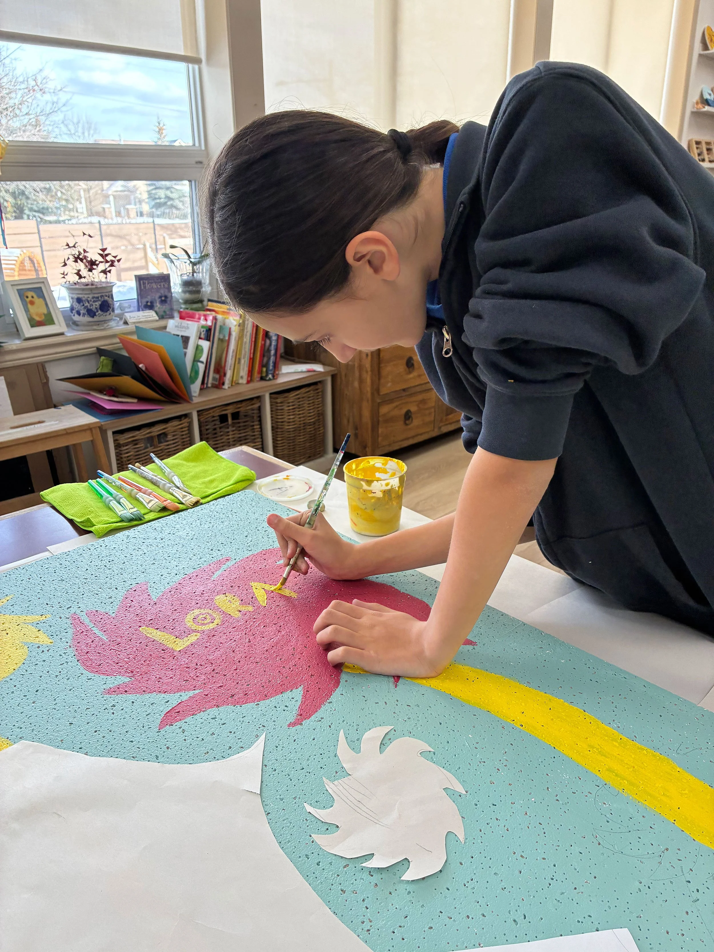A person paints a colorful model of a flamingo with the word 'LOVE' in yellow on its body, on a speckled blue surface with white paper cutouts of a flamingo and another bird nearby.