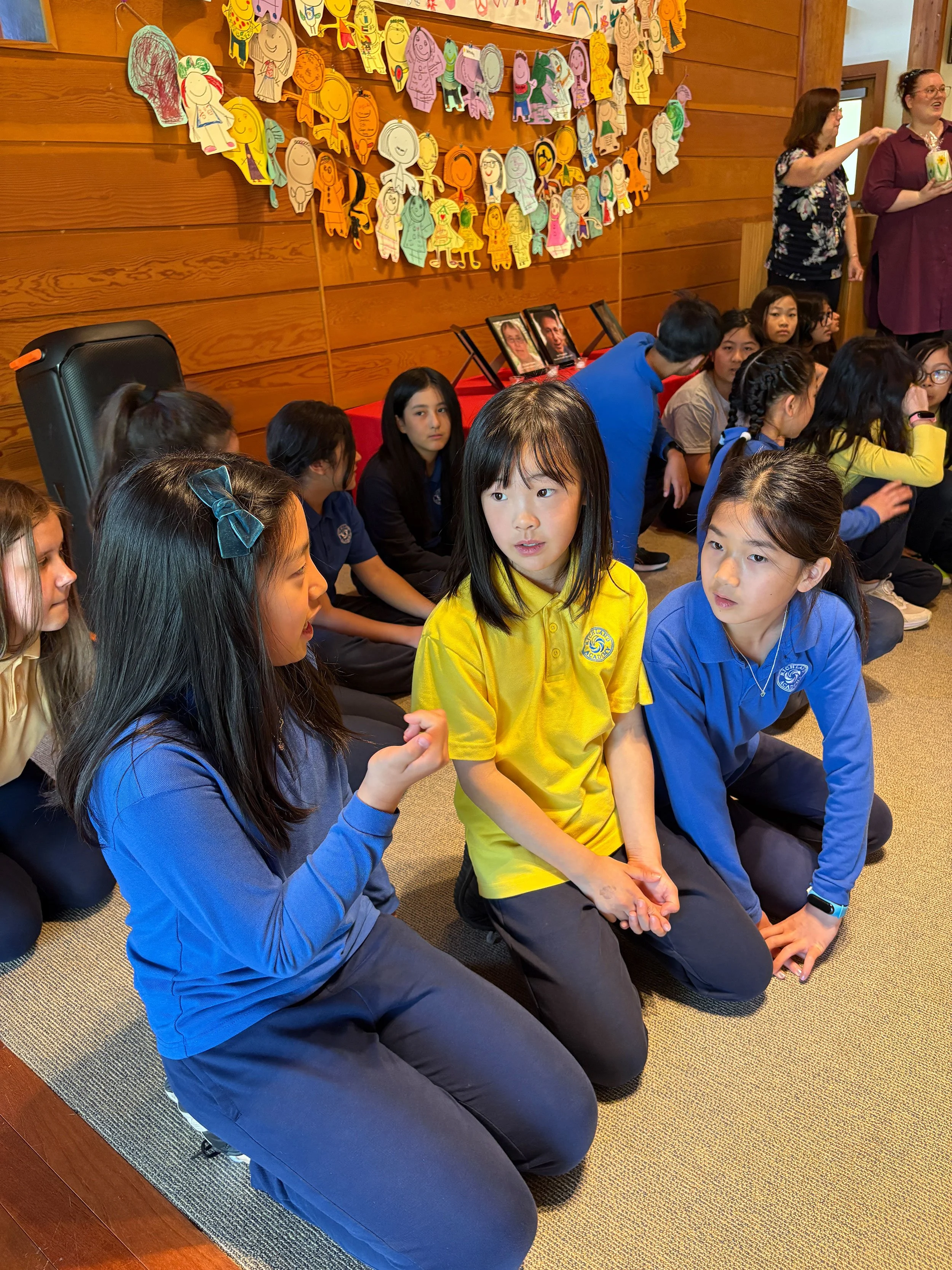 A group of children sitting cross-legged on the floor, engaging in a conversation, with two women standing in the background near a display of colorful drawings and framed photographs.