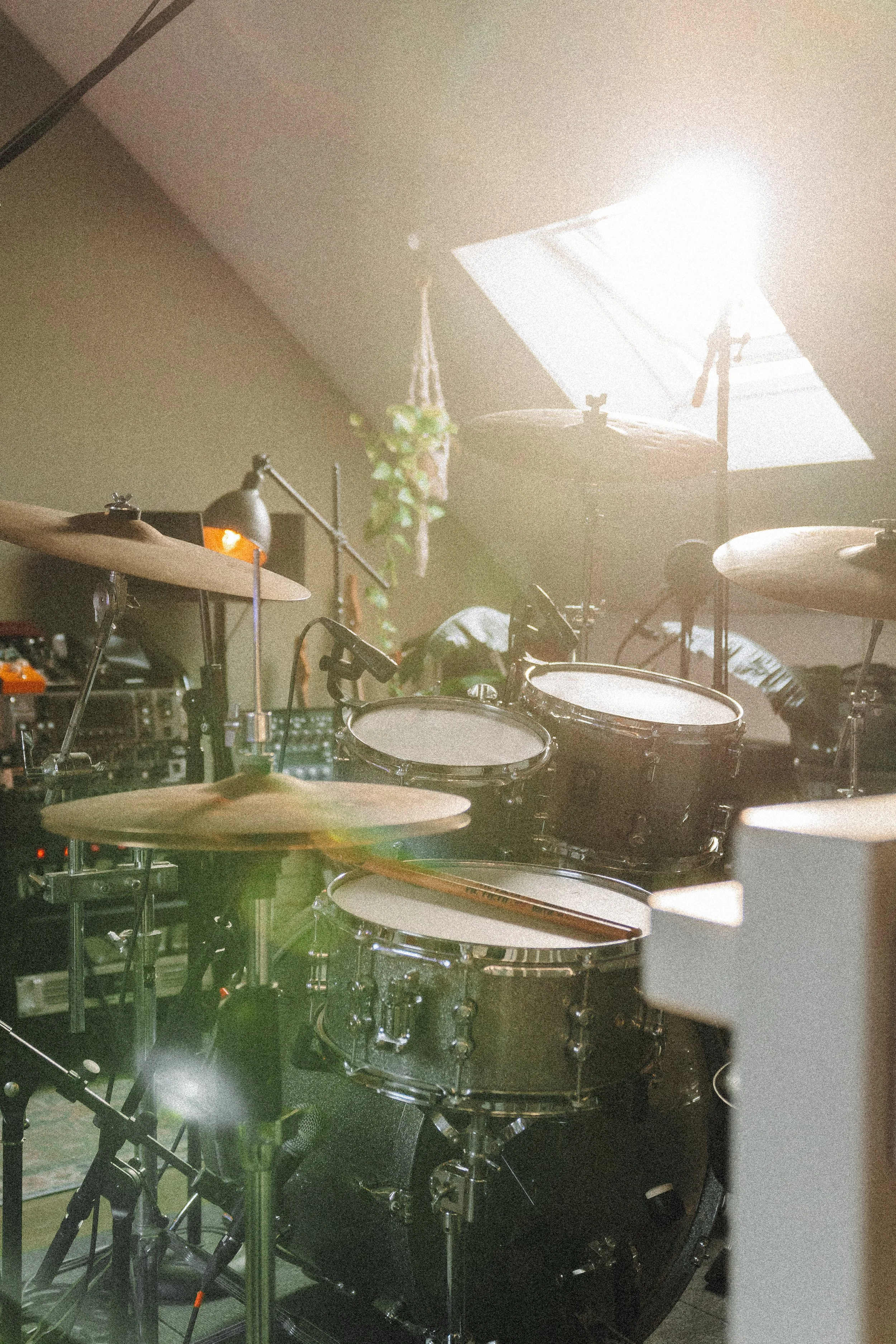 Binnenaanzicht van een muziekstudio met een drumstel, microfoons en andere audioapparatuur. Een persoon zit te spelen op de achtergrond.