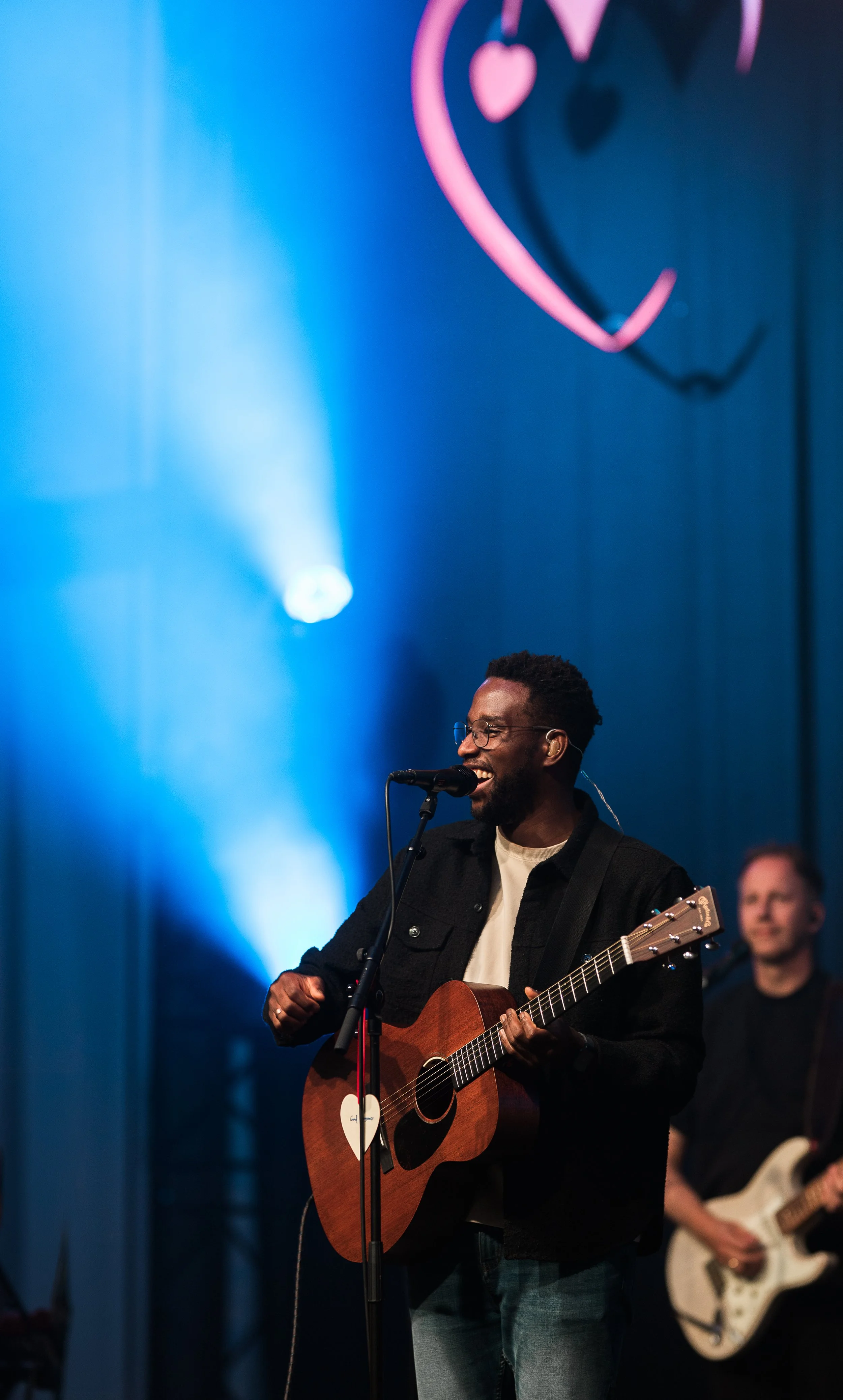 Een man die glimlachend zingt en gitaar speelt tijdens een liveoptreden op een podium met blauwe en roze verlichting, met andere muzikanten op de achtergrond.