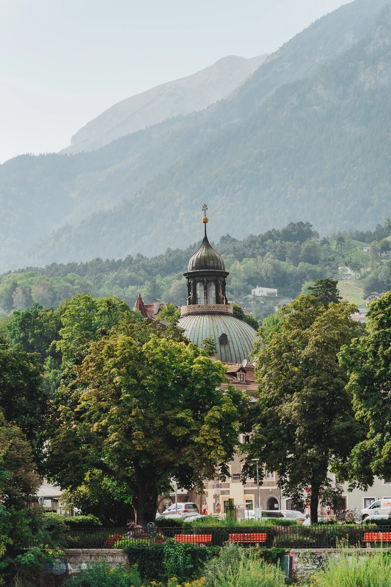 Een dorp met een kerk met een koepel en kruis op de top, omringd door groene bomen, met bergen op de achtergrond en een plein met auto's en mensen.