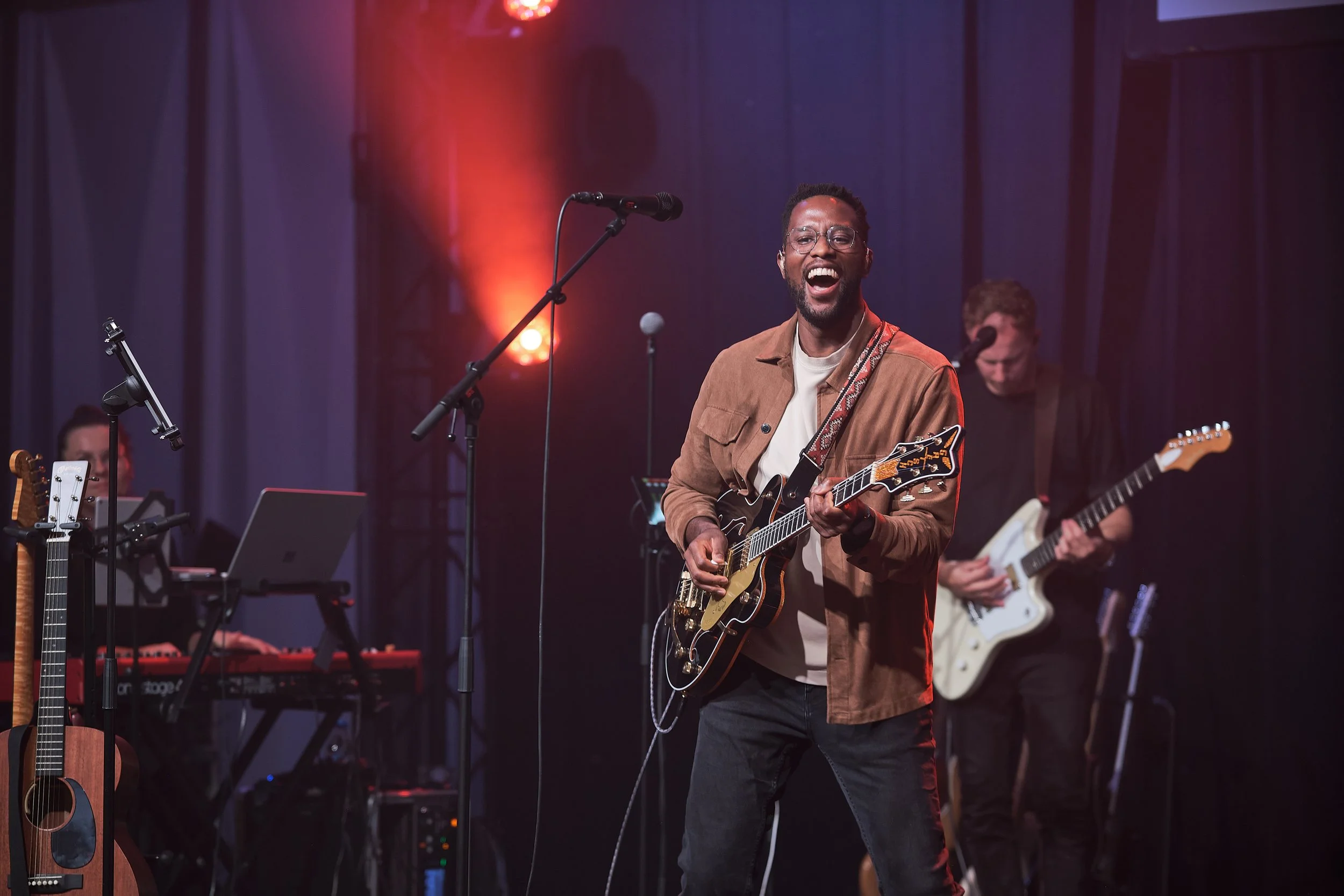 Anthony Trustfull speelt gitaar op podium van Heart2Heart met muzikanten op de achtergrond.