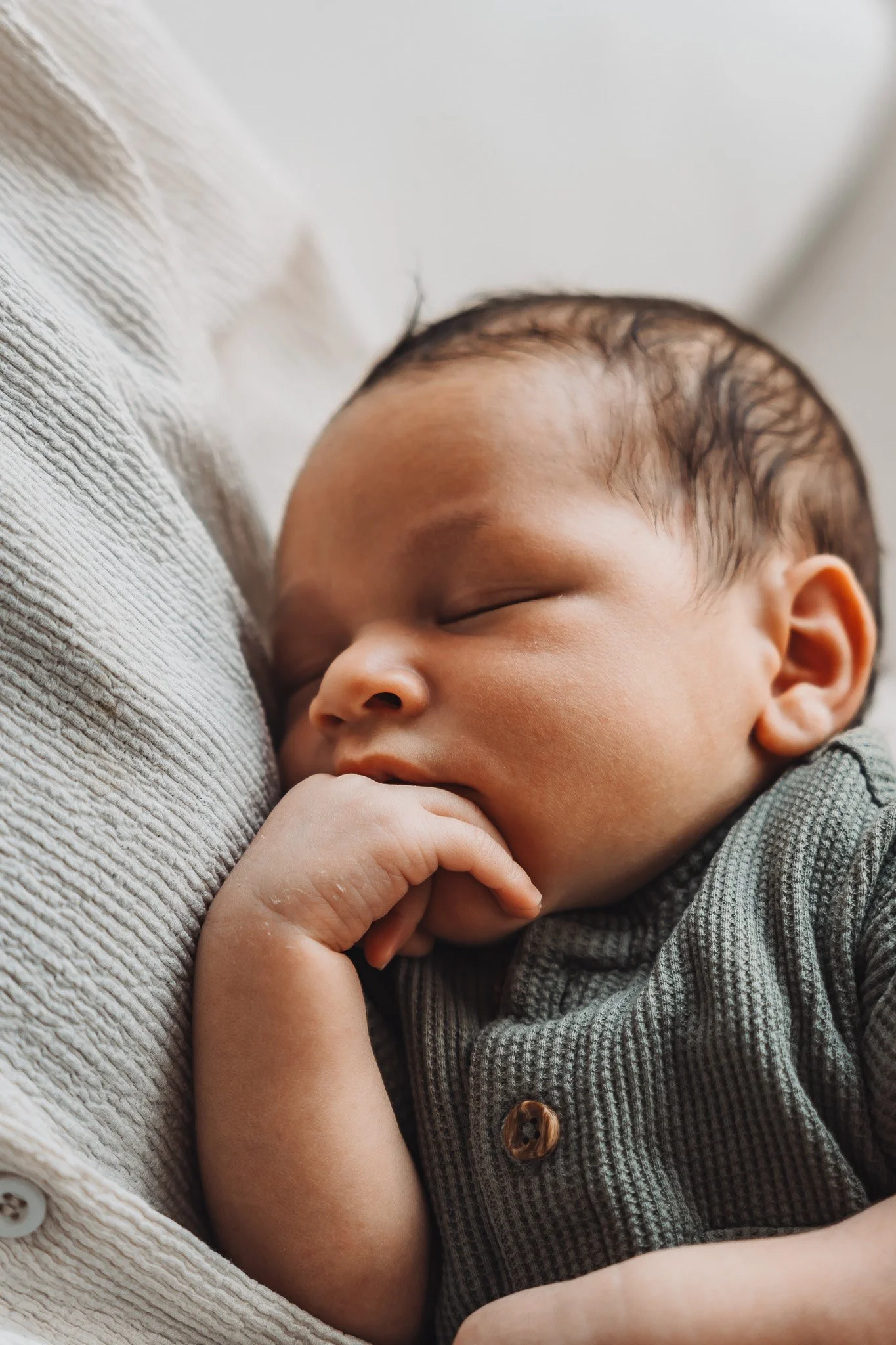 Close-up van een slapende baby met donkere haar, gekleed in een grijs geruit shirt, rustend op een witte, ribbelige stof.