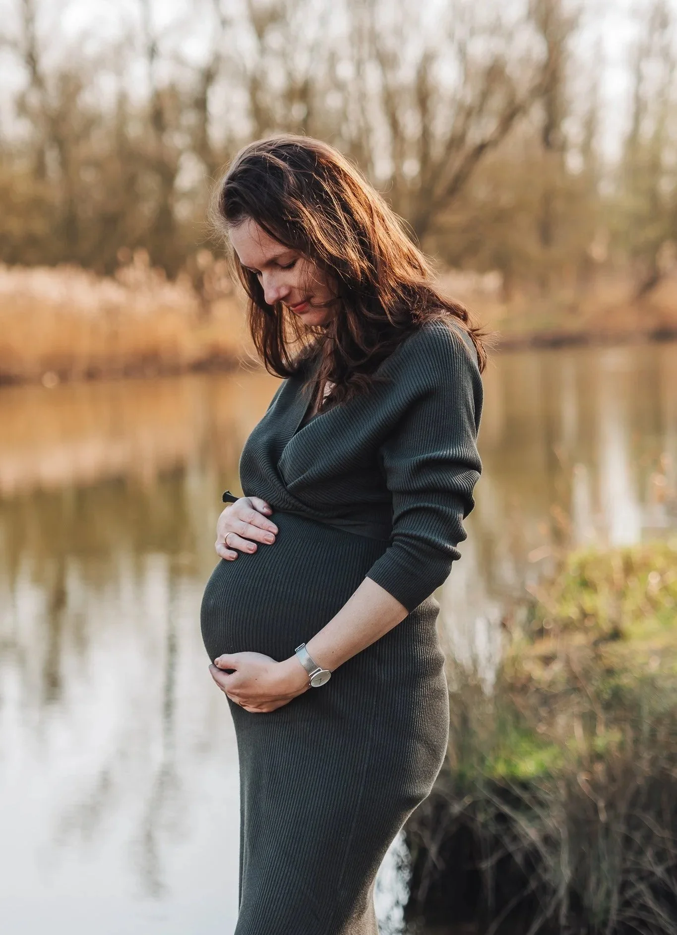 Een zwangere vrouw met lang bruin haar in een groene jurk staat naast een rivier, haar handen rusten op haar buik en ze kijkt naar beneden in een natuurlijke omgeving met bomen en water.