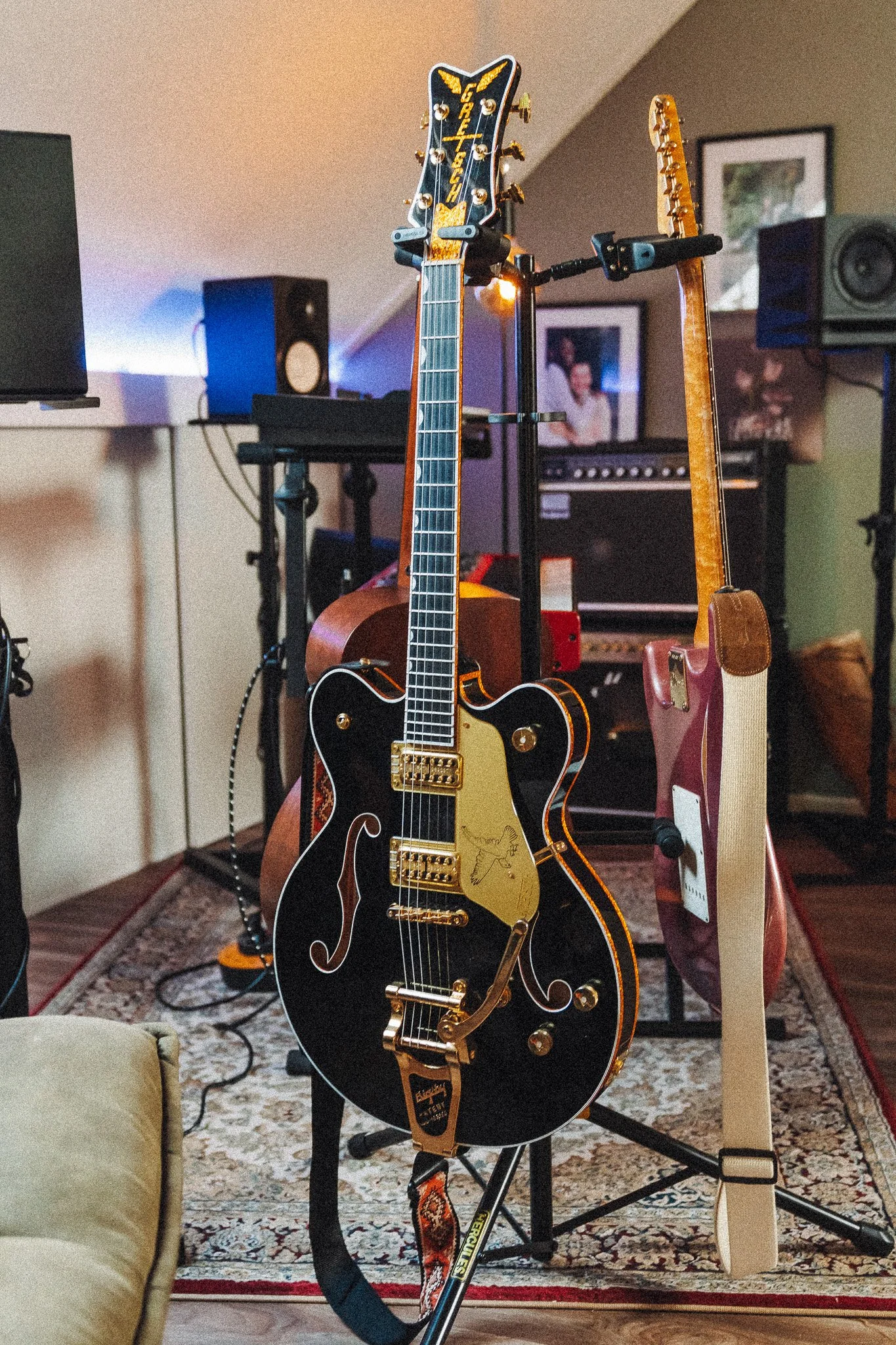 Een opname van een muziekstudio met twee elektrische gitaren en een akoestische gitaar op een standaard, omringd door versterkers en audioapparatuur.