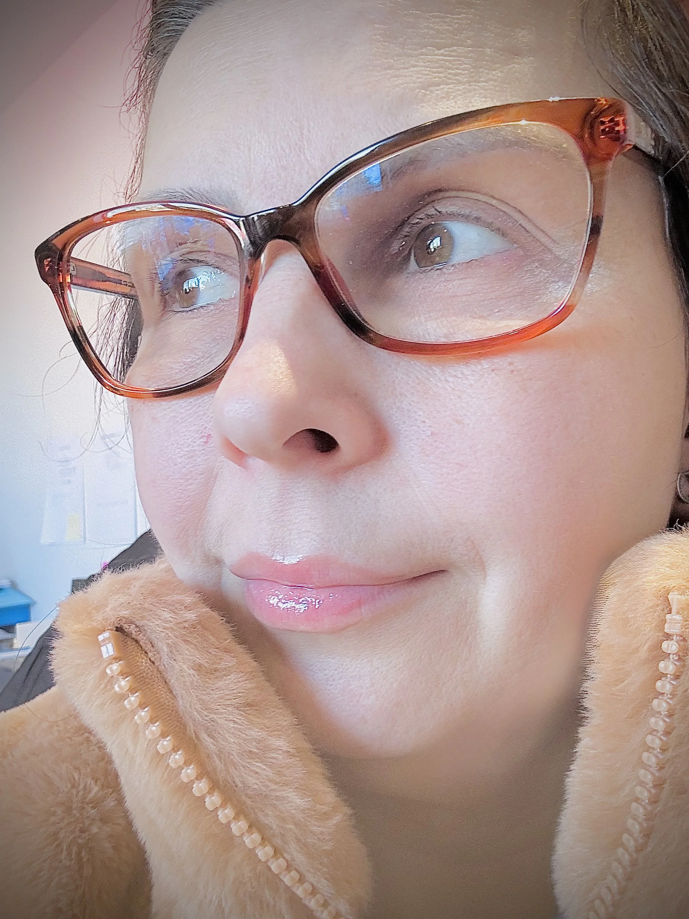 Close-up of a woman with glasses, wearing a tan furry jacket with pearl decorations, looking slightly away from the camera.