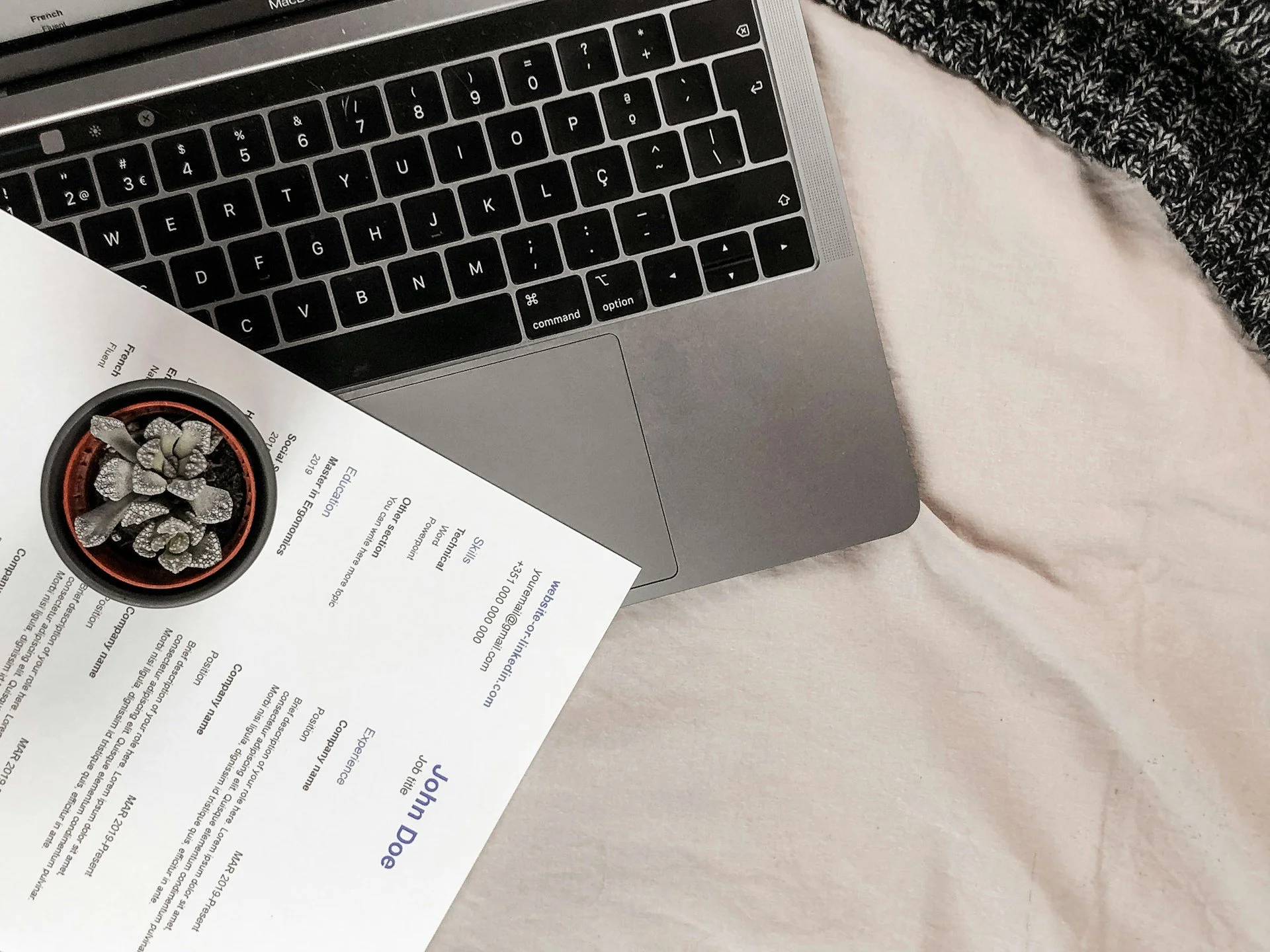 Close-up of a silver laptop keyboard on a light-colored surface, with a partially visible printed document featuring a small potted cactus plant.