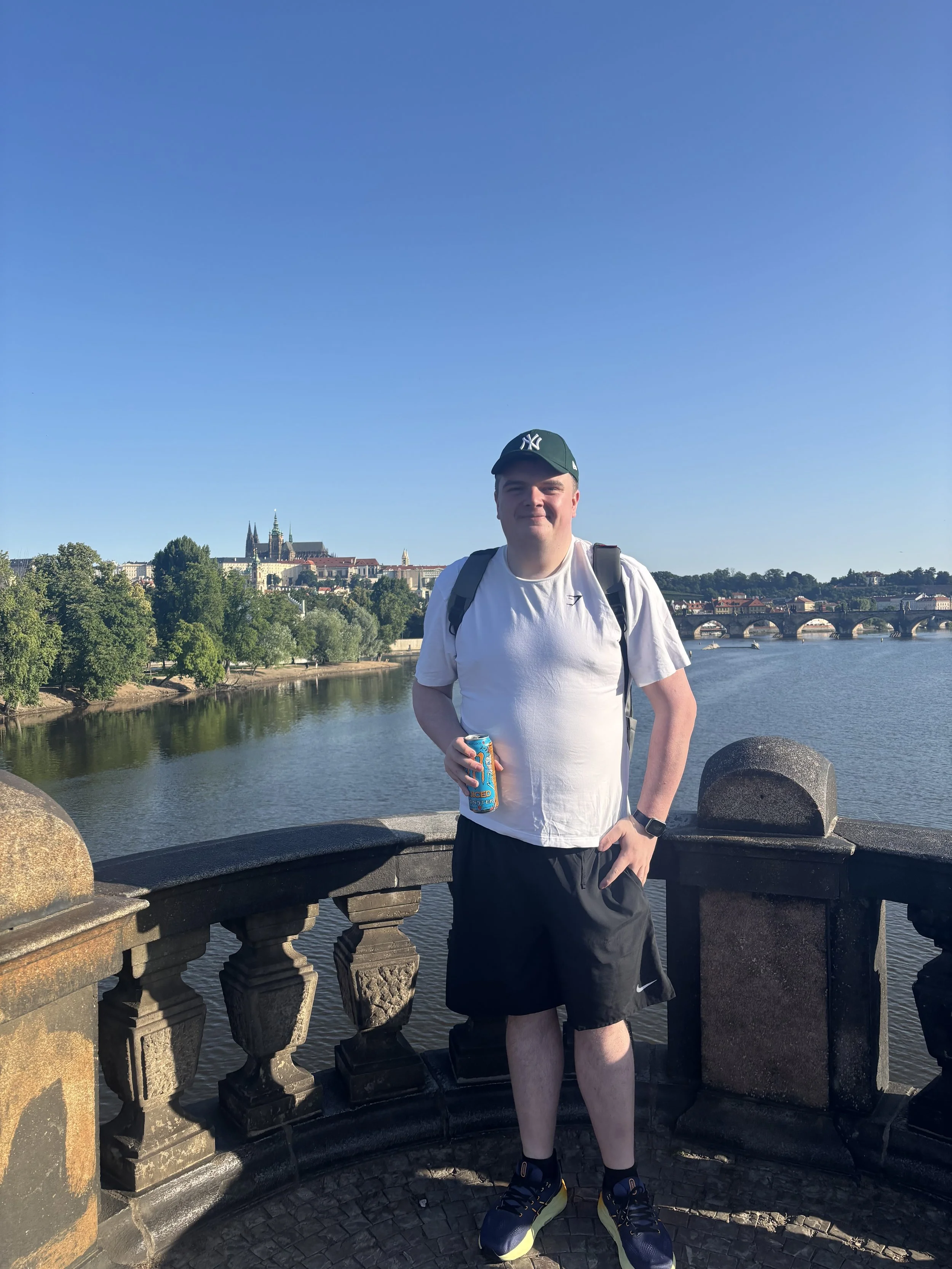 A young man standing on a bridge over a river, holding a can of soda, with a cityscape and a cathedral in the background under a clear blue sky.
