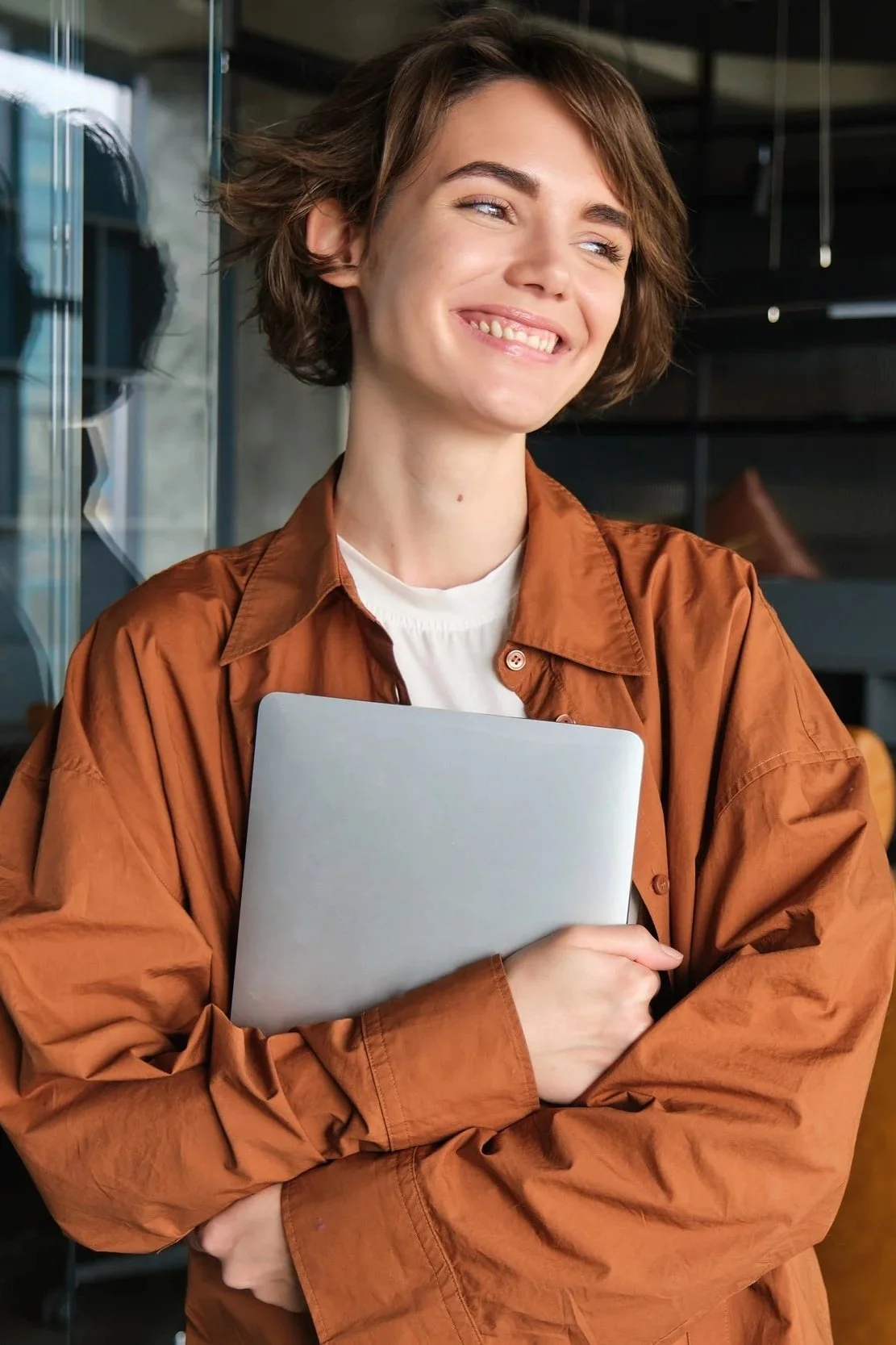 A young woman with short brown hair, smiling, holding a silver laptop in an indoor setting with glass and modern decor.