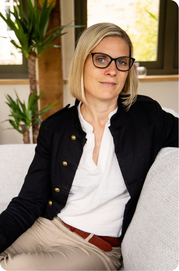 A woman with blond hair and glasses sitting on a light-colored couch in a room with large windows and indoor plants.
