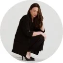 Woman with long brown hair wearing black clothing, crouching against a plain white background.