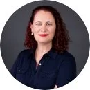 A woman with curly red hair wearing a dark blue shirt against a dark background.