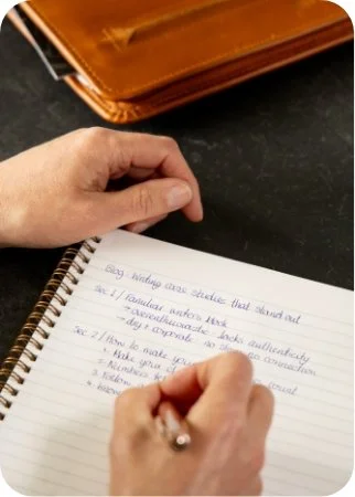 A person writes in a spiral notebook with a silver pen, with a brown leather binder in the background on a black surface.