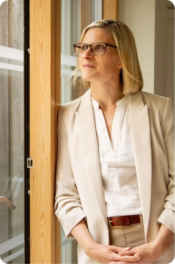 A woman with blonde hair and glasses looking out a window, dressed in a light-colored blazer and white shirt, standing indoors near a wooden door or wall.