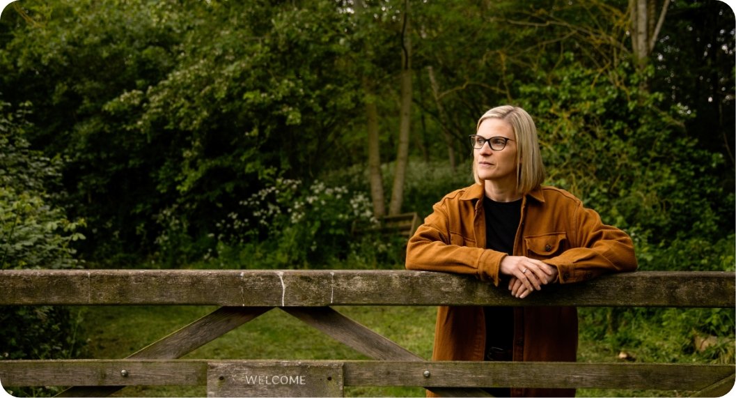 A woman with glasses and gray hair leaning on a wooden bridge railing in a lush green park.