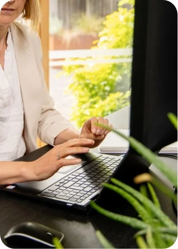 A woman sitting at a desk using a computer with a window showing greenery outside.