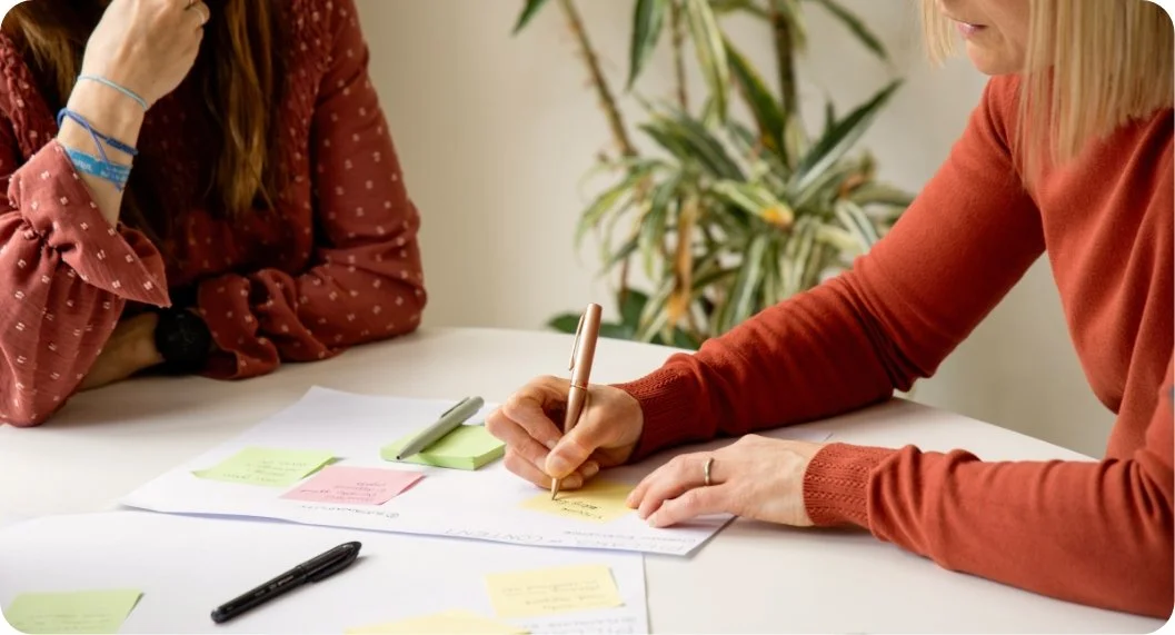 Two women sitting at a white table with papers, sticky notes, and pens, engaged in a discussion or brainstorming session.