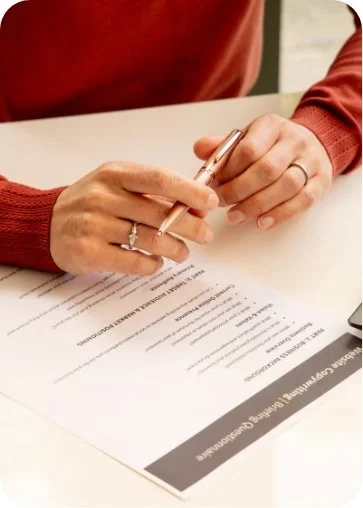Person in a red sweater signing a document with a pen on a desk.