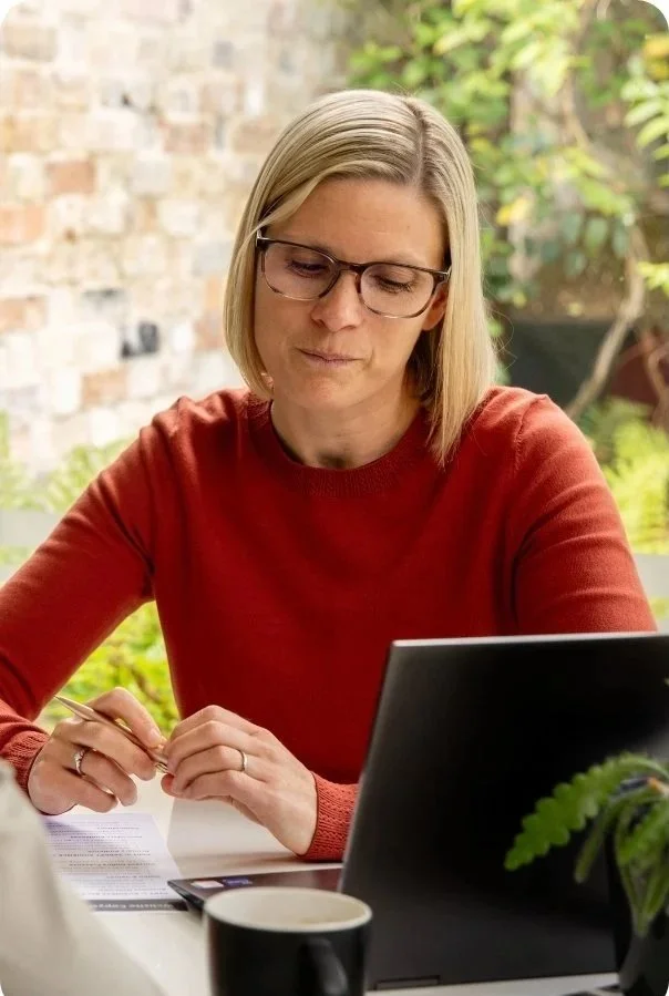 Woman with blonde hair and glasses wearing a red sweater working at a table outdoors with a laptop, notebook, pen, a cup, and plants in the background.