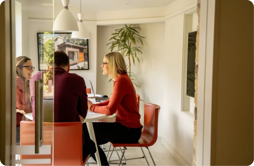 Three people in a meeting room, two women and one man, sitting at a table with laptops, engaged in conversation.