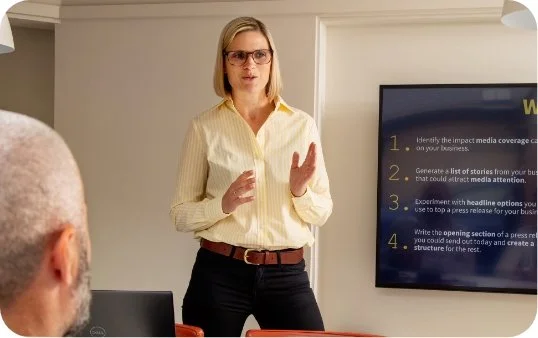 Woman giving a presentation in a conference room with a large screen displaying a presentation slide.