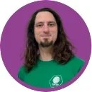 A man with long dark hair and a beard, wearing a green shirt, standing in front of a purple background.