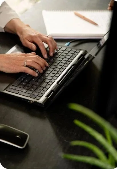 Person typing on a laptop keyboard with a notebook and pencil nearby.