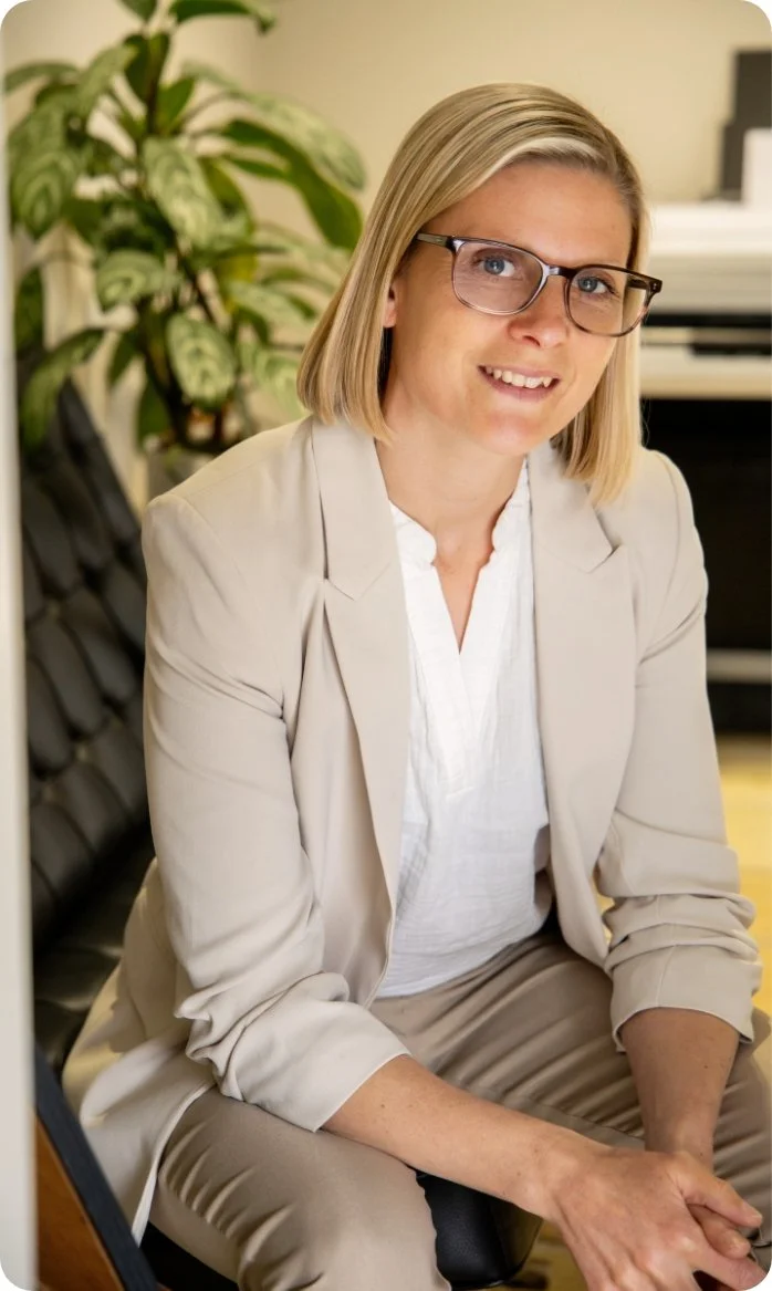A woman with blonde hair and glasses sitting on a black chair in an office, smiling, with a green plant and a piano in the background.