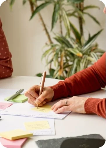 Person writing with a gold pen on a notepad surrounded by colorful sticky notes and papers, with green plants in the background.