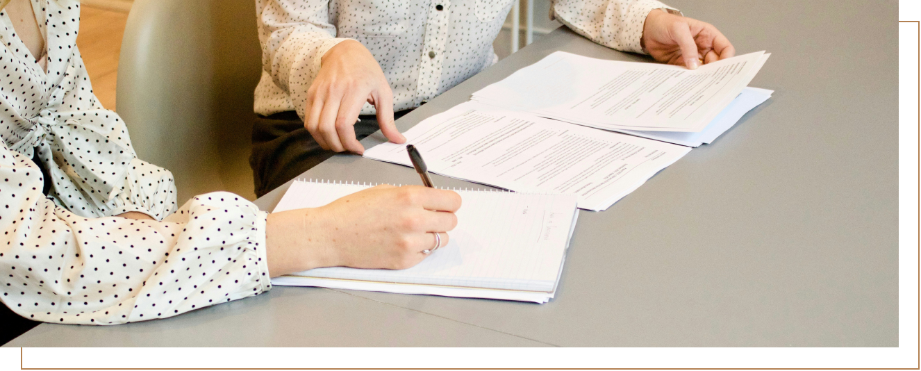 Two people sitting at a table with documents, one is writing on a notepad, the other is pointing at the papers.