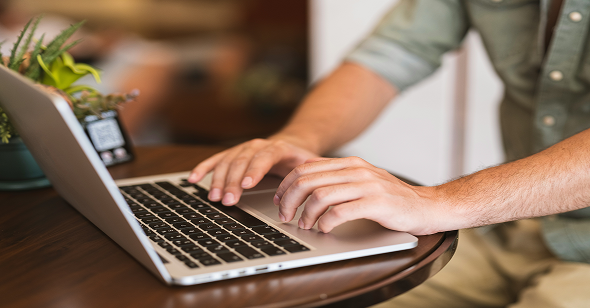 Person working on a laptop at a wooden table with a potted plant and a phone nearby.