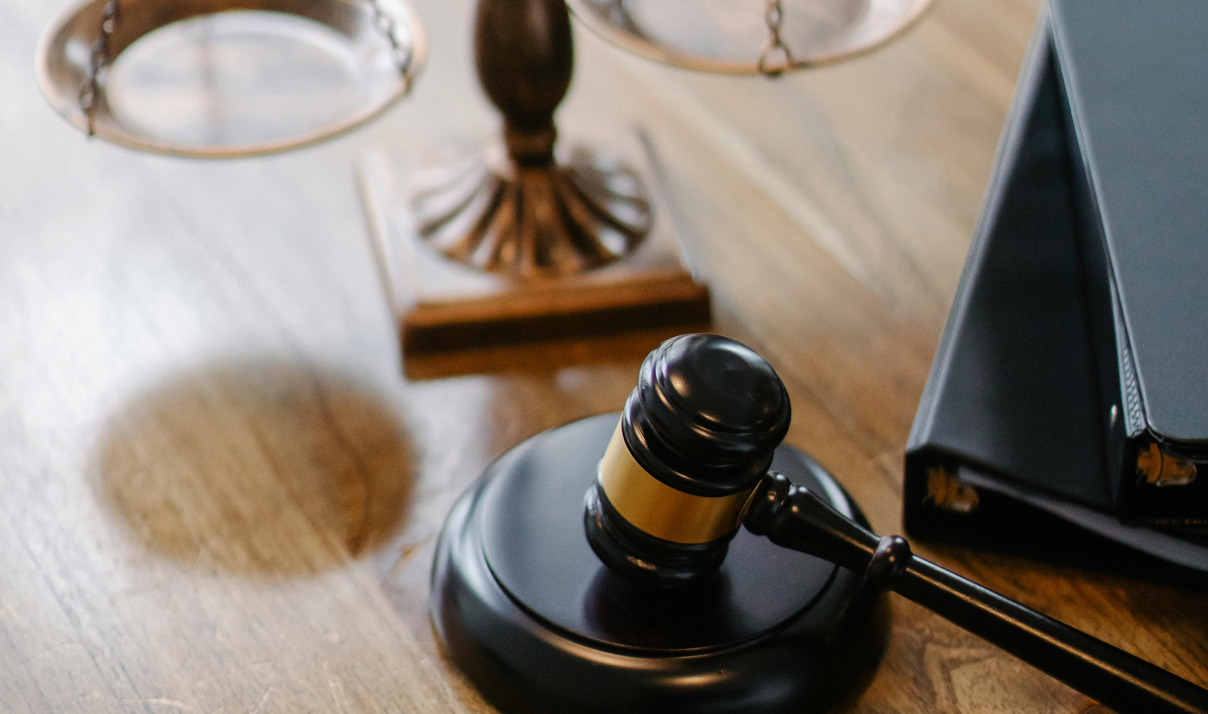 A judge's gavel resting on a wooden desk next to a legal scale and a black binder.