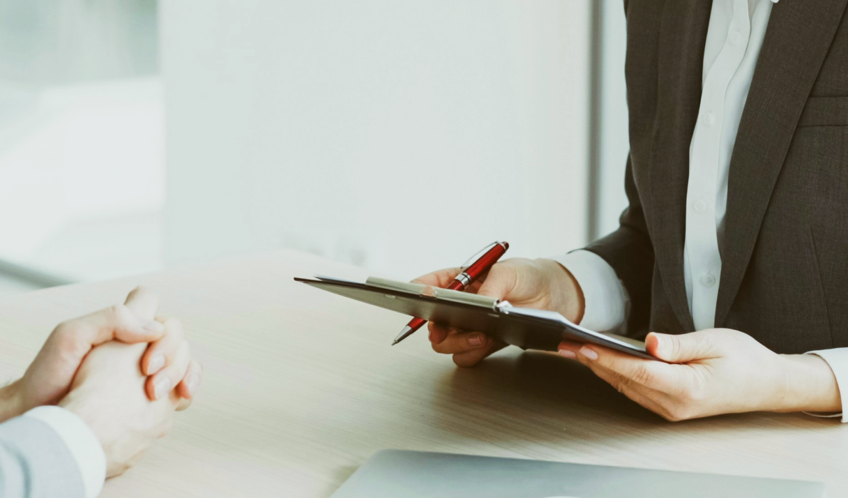 Business meeting with one person holding a clipboard and pen, another person's hands clasped on the table, in a professional setting.