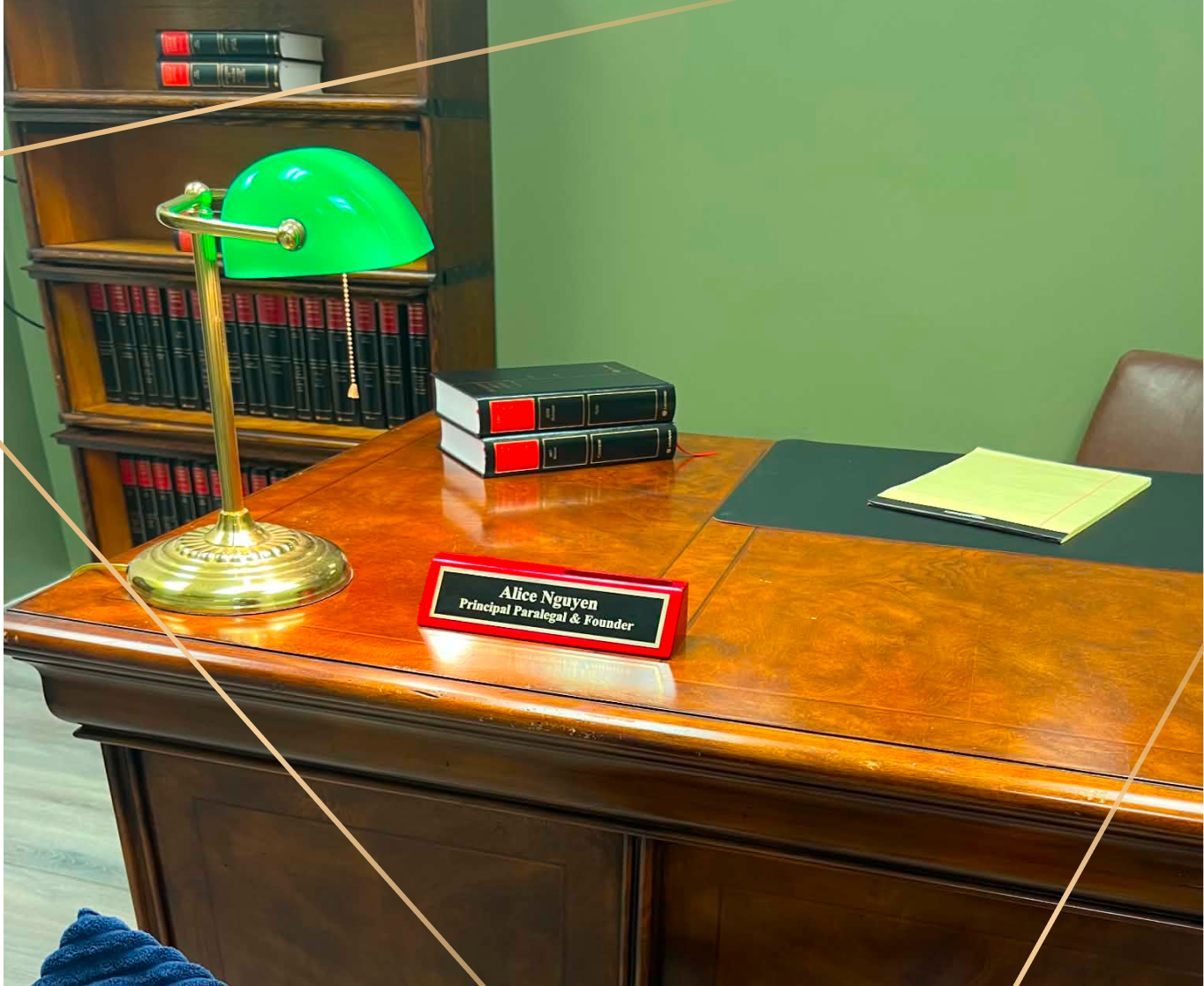 An office desk with a green banker lamp, three stacked black law books, a yellow notepad, a black folder, and a red and black nameplate that reads 'Alice Nguyen, Principal Paralegal & Founder.' In the background, a wooden bookshelf with red and black legal books.