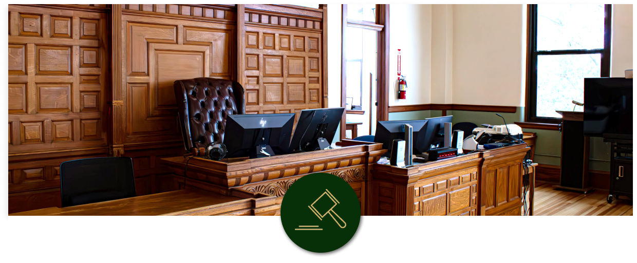 Interior of a courtroom with wooden walls, a judge's bench with three computer monitors, a leather chair behind the bench, and windows letting in natural light.