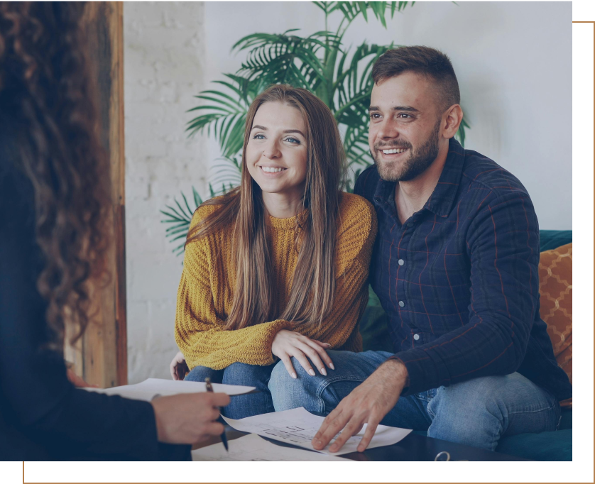 A couple sitting on a couch during a consultation with a therapist. The woman has long, straight hair and is wearing a mustard yellow sweater. The man has short hair and a beard, wearing a dark blue plaid shirt. They are smiling and looking at the therapist, who is holding a notepad and pen.