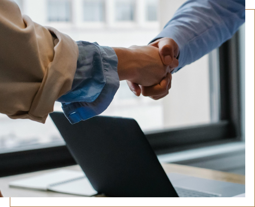 Two people shaking hands in an office setting with a laptop and a notebook on the table.
