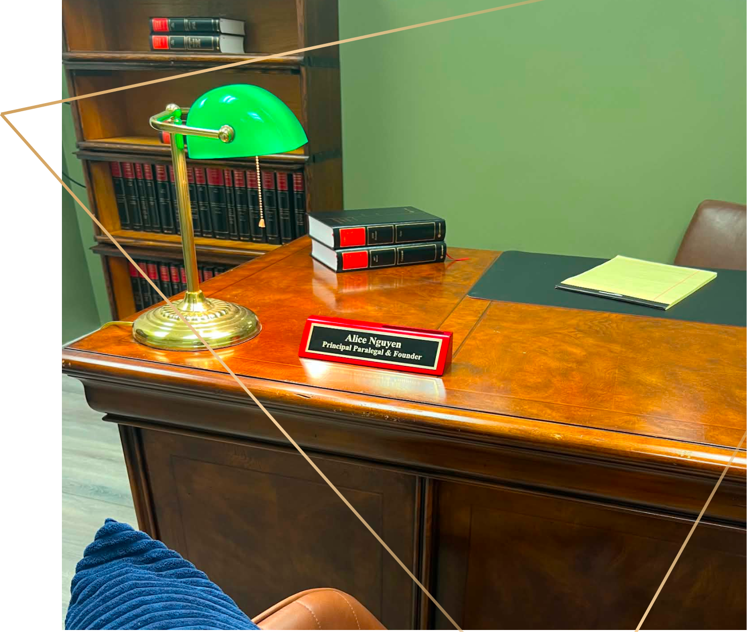 A polished wooden office desk with a green banker’s lamp, a stack of three black and red legal books, a nameplate that reads 'Alice Nguyen, Principal Paralegal & Founder,' a yellow legal pad with a pen, a black desk pad, and a leather chair.